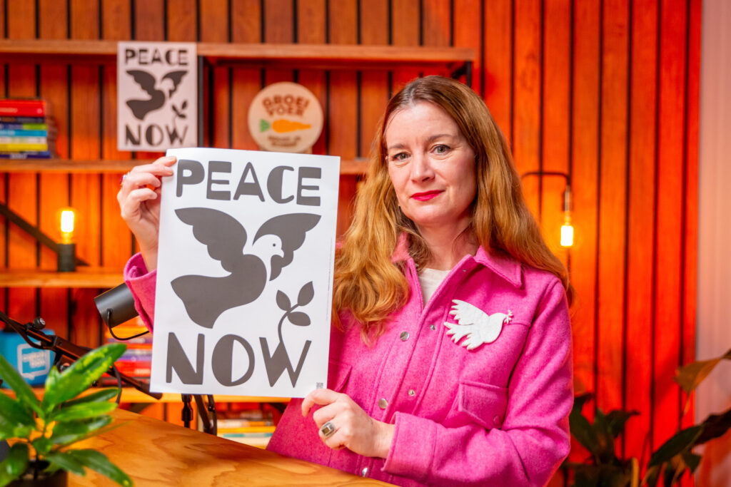 Vrouw in roze jas houdt een poster vast met de tekst "Peace Now" en een afbeelding van een vredesduif, in een kleurrijke podcaststudio.