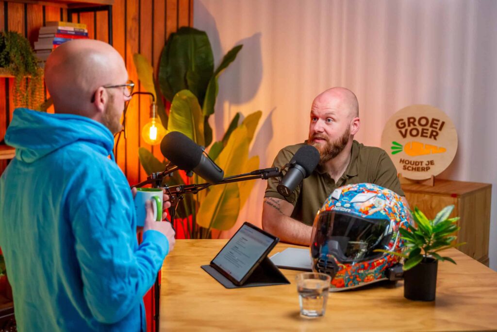 Twee mannen in gesprek tijdens een opname van de Groeivoer-podcast, met microfoons, een kleurrijke motorhelm en planten op tafel.
