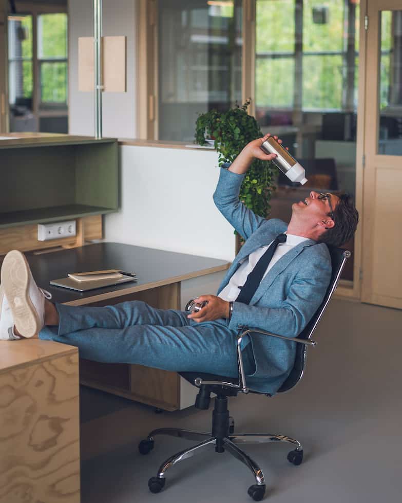 Man in een blauw pak drinkt uit een Dopper-fles terwijl hij achteroverleunend met zijn voeten op het bureau zit.