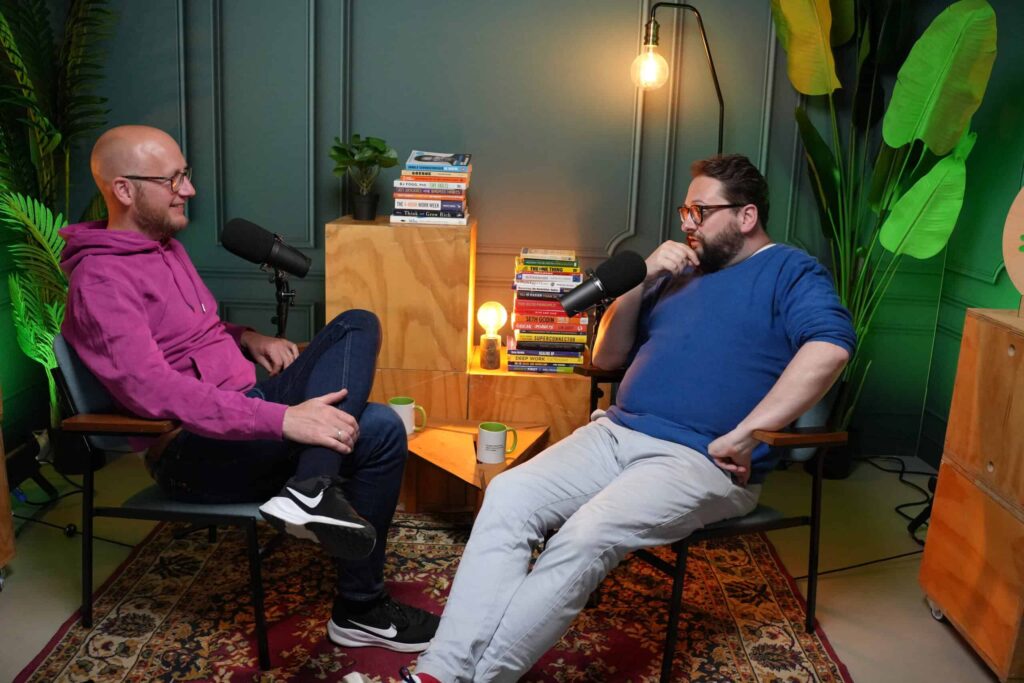 Twee mannen voeren een gesprek in een gezellige podcaststudio met microfoons, boeken en sfeerverlichting.