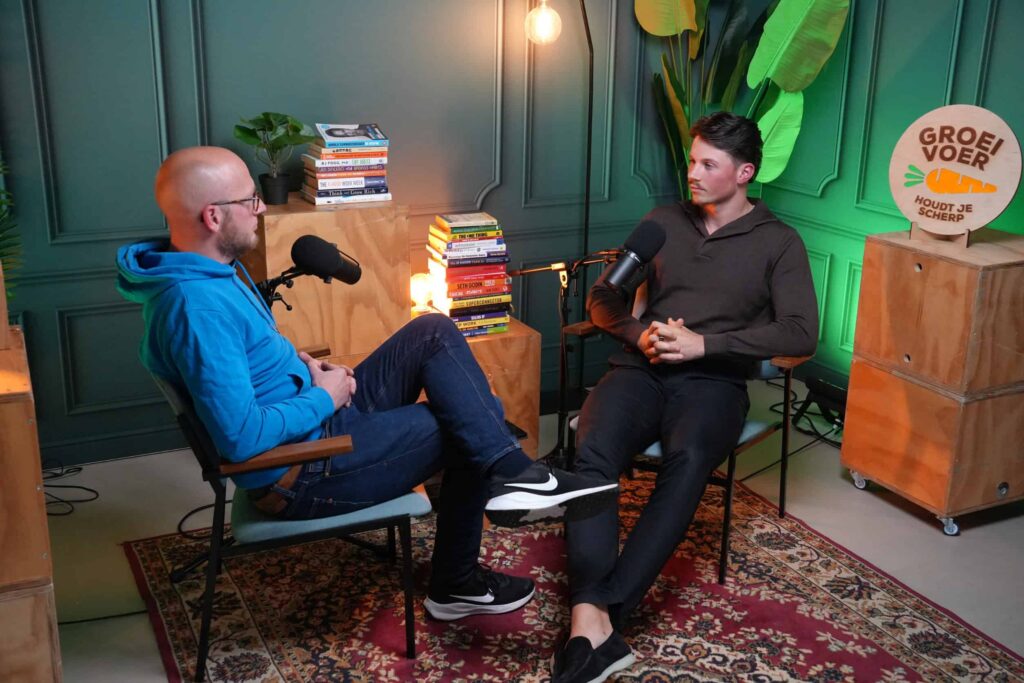 Twee mannen zitten tegenover elkaar in een podcaststudio met microfoons en stapels boeken tussen hen in.