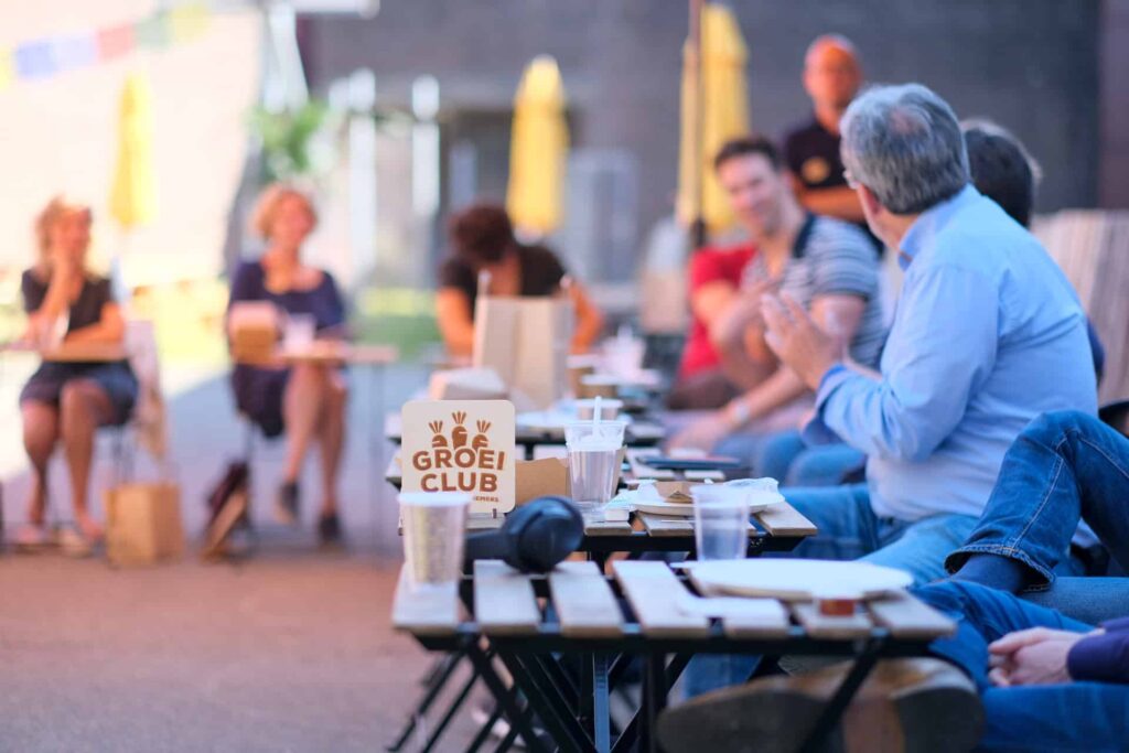 Mensen zitten buiten aan tafeltjes tijdens een bijeenkomst van de Groei Club.