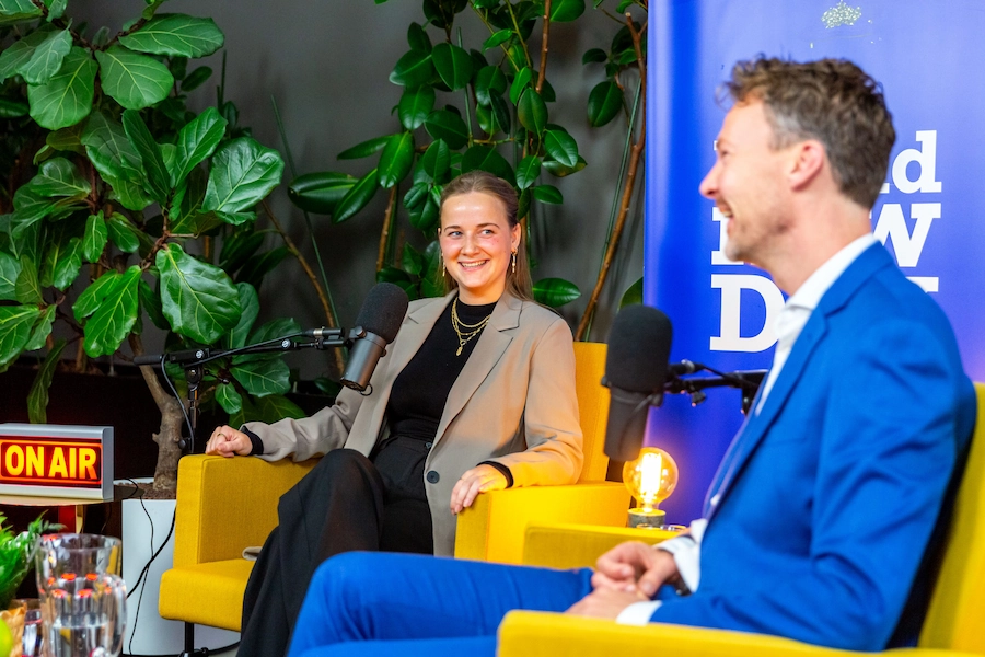 Twee mensen zitten in gele stoelen en praten in microfoons tijdens een opname van de podcast "Brand New Day".