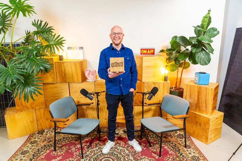 Man in een blauwe blouse houdt een bord met het logo "EAR EAR" vast in een kleurrijk ingerichte podcaststudio met microfoons en stoelen.