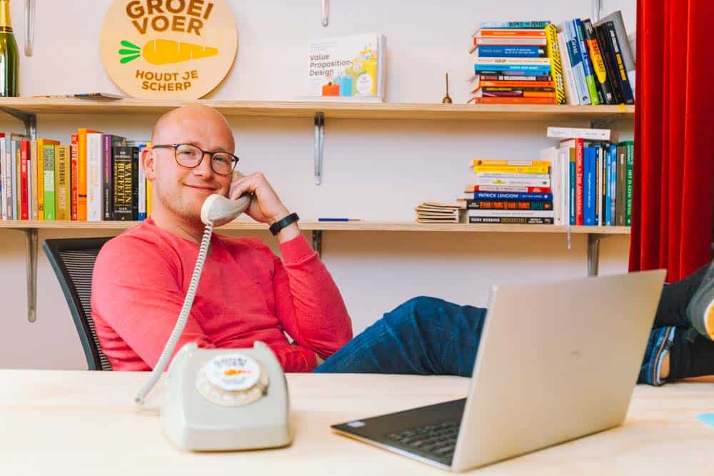 Gerhard te Velde zit ontspannen met een ouderwetse telefoon aan zijn oor in een kleurrijk kantoor met boekenplanken op de achtergrond.