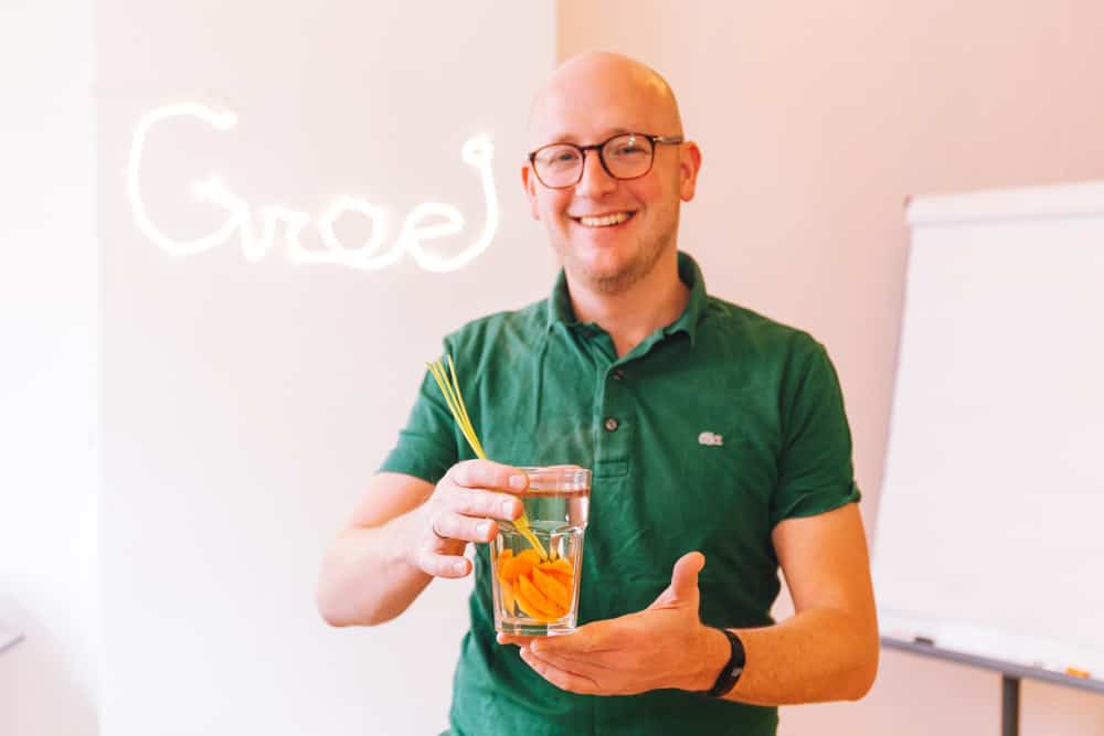 Gerhard te Velde houdt een glas met water en sinaasappelpartjes vast voor een flip-over met het woord "Groei" op de achtergrond.