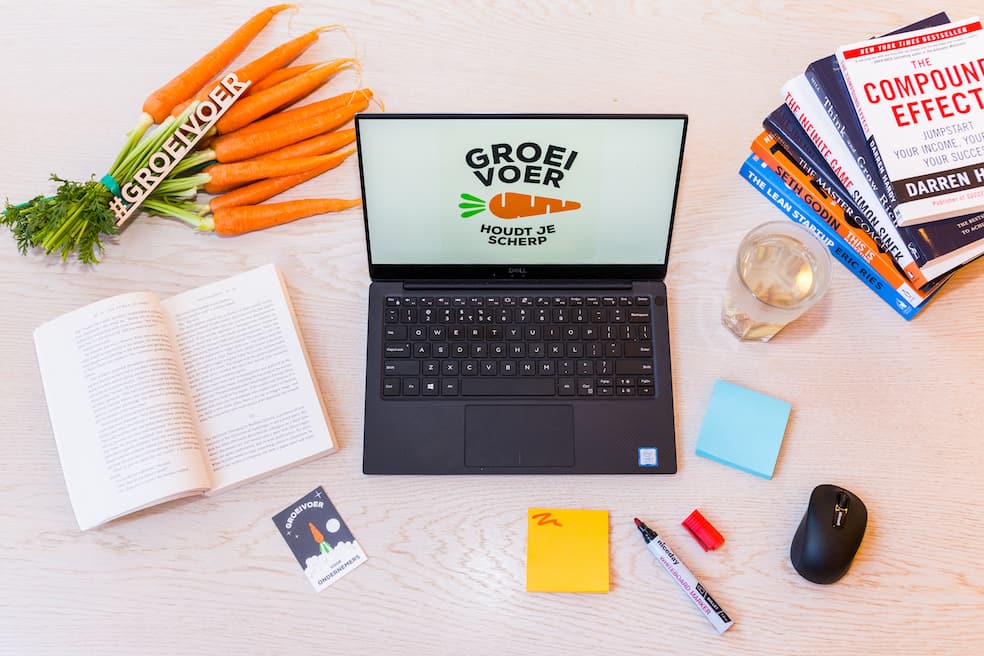 Werkstation van Groeivoer met laptop, boeken, wortels en kantoorbenodigdheden op een houten bureau.