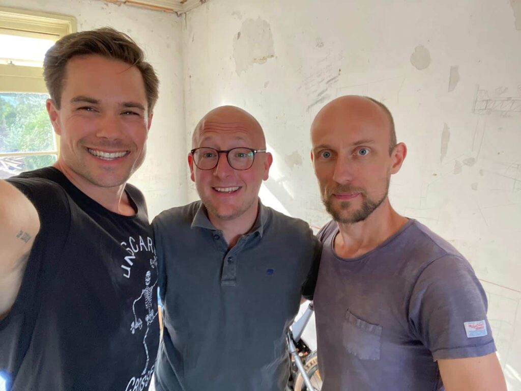 Drie lachende mannen poseren samen voor een selfie in een lichte, onafgewerkte kamer.