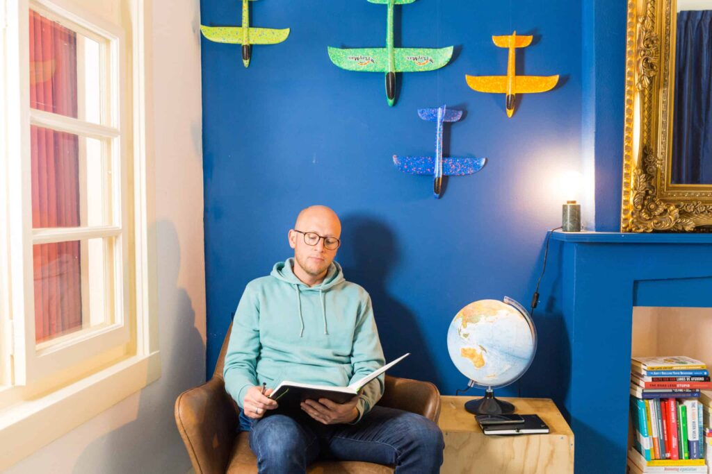 Man in een fauteuil leest een boek in een kleurrijke kamer met wereldbol, boeken en modelvliegtuigjes aan de muur.