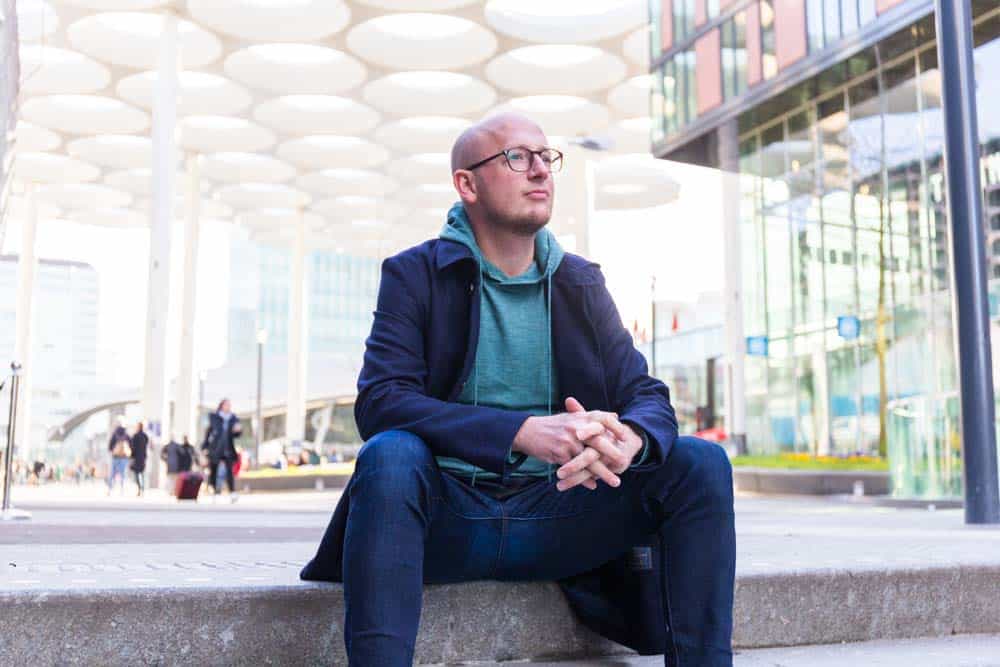 Man met bril zit op een betonnen rand bij het Stadskantoor in Utrecht, onder de markante ronde overkapping van het Stationsplein.