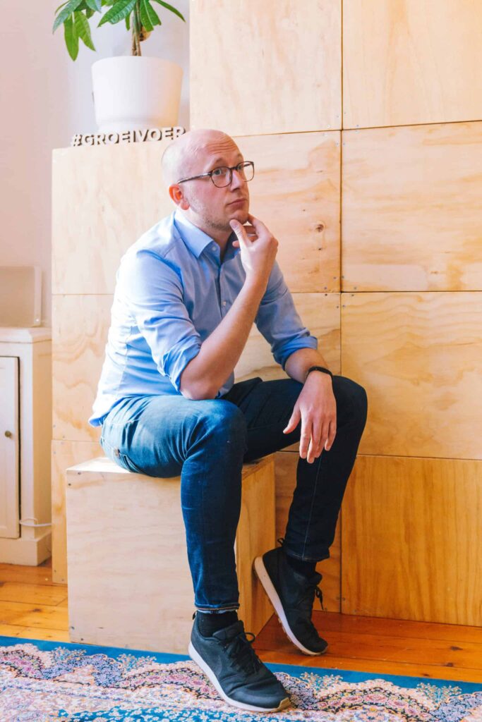 Man in blauwe blouse zit peinzend op een houten kist in een modern interieur met de hashtag #GROEIVOER op de achtergrond.