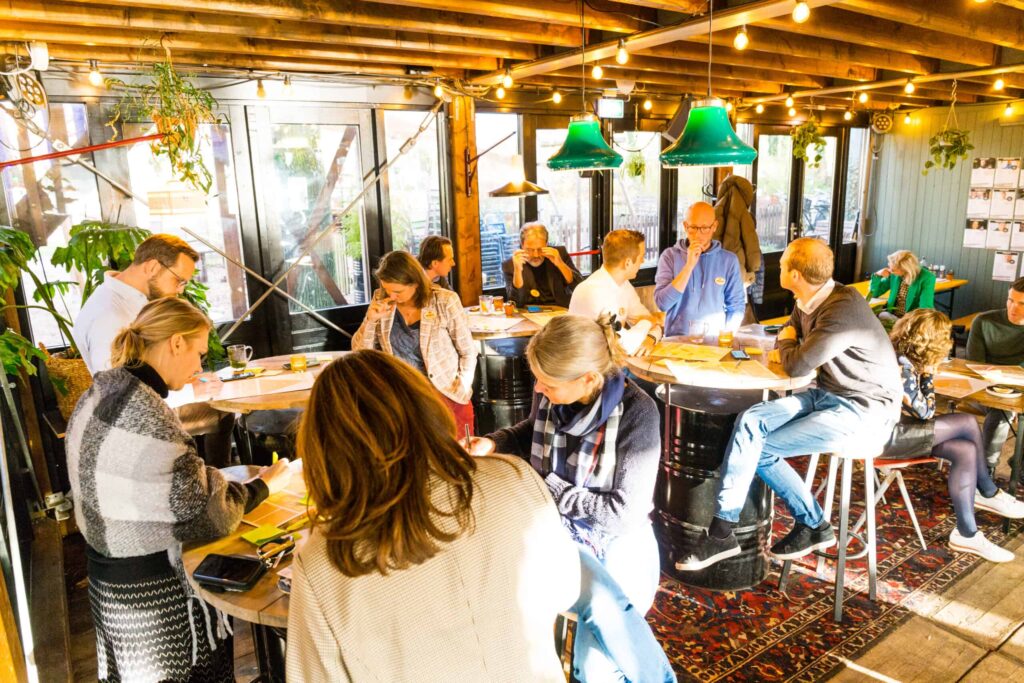 Groep mensen werkt samen aan tafels in een zonnige, gezellig ingerichte ruimte met houten plafond en hanglampen.