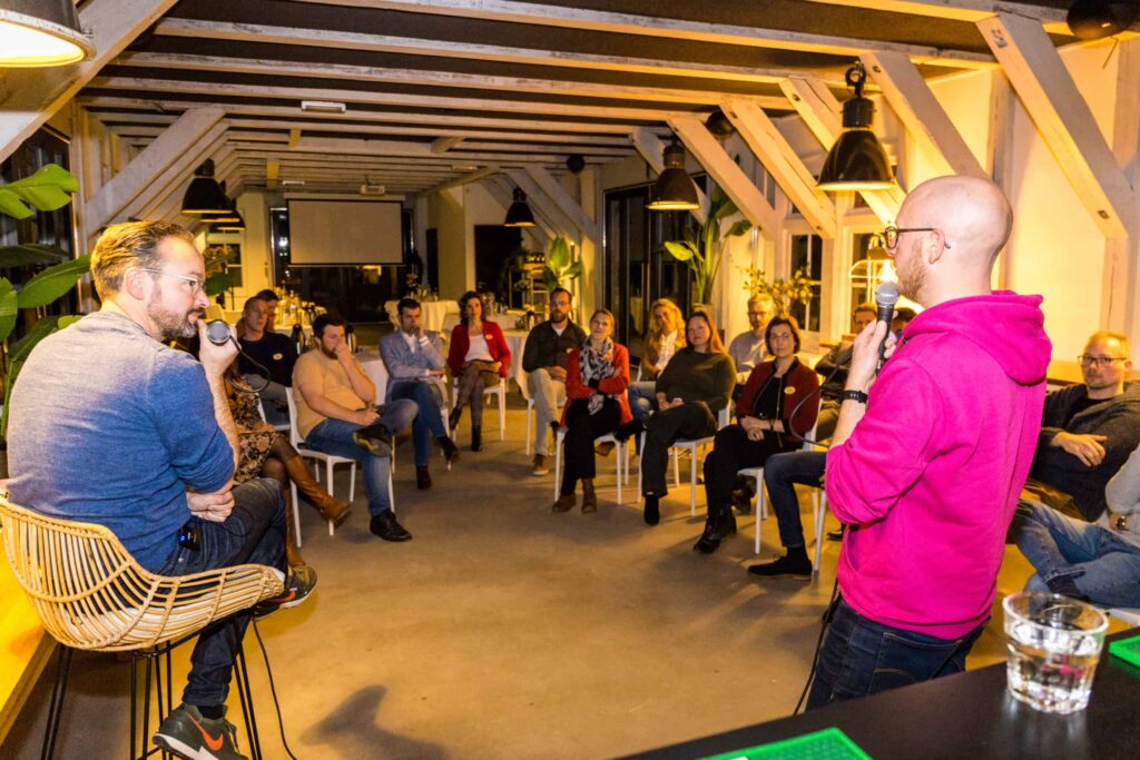 Een spreker in een felroze trui praat tot een aandachtig luisterend publiek tijdens een kleinschalig evenement in een sfeervolle ruimte.
