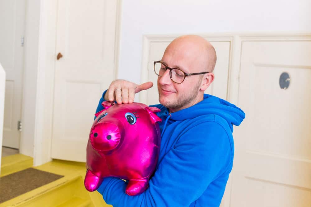 Man in blauwe trui houdt een glimmend roze spaarvarken vast en glimlacht tevreden.