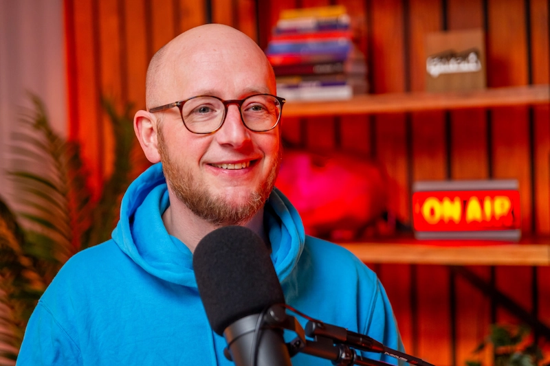 Man met bril en blauwe hoodie spreekt in microfoon tijdens een podcastopname in een kleurrijke studio met "ON AIR"-bord op de achtergrond.