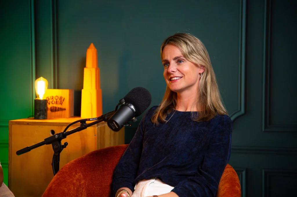 Blonde vrouw als podcastgast in studio met microfoon en sfeerverlichting.