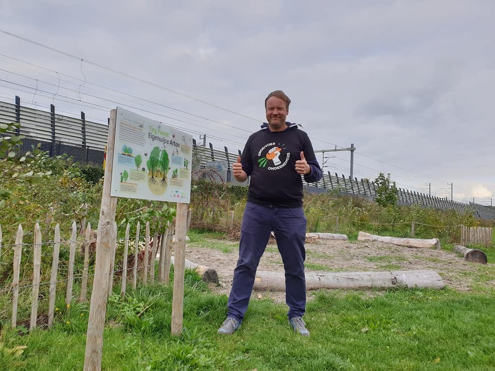 Jannes Herman Mostert van The Tree Party geeft een duim omhoog bij een Tiny Forest naast een informatiebord, terwijl hij een Groeivoer-shirt draagt.