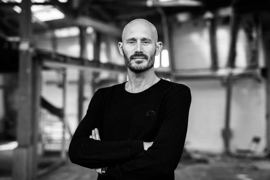 Zwart-wit portret van spreker Klaas Boomsma met gekruiste armen in een industriële omgeving.