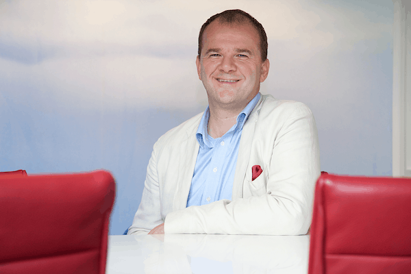 Marc Wesselink zittend aan een witte vergadertafel met rode stoelen, gekleed in een licht colbert en blauw overhemd.