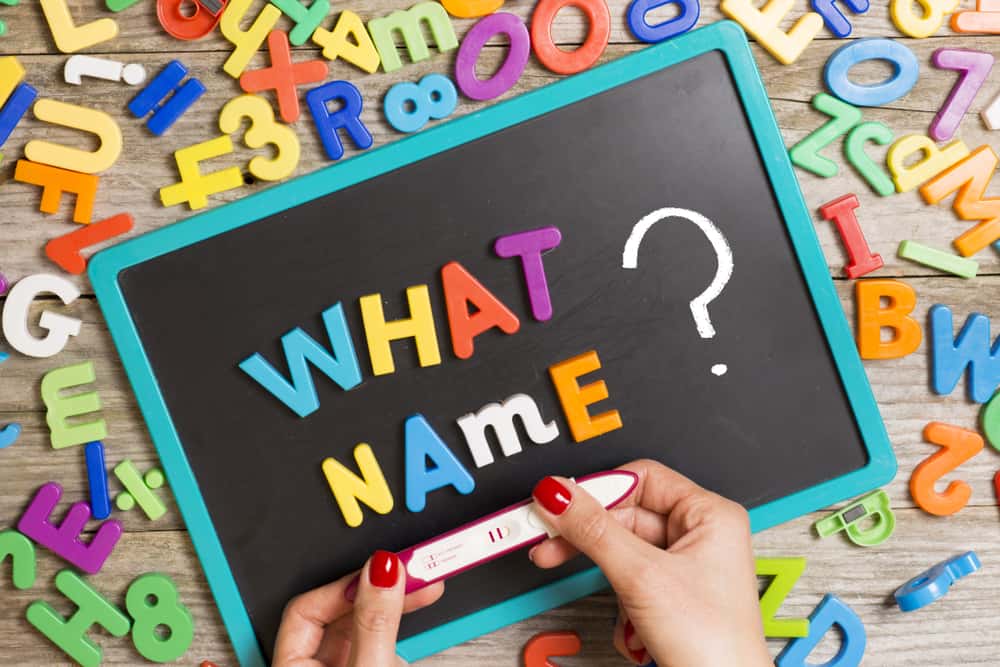Een schoolbord met kleurrijke letters die 'WHAT NAME?' spellen, omringd door losse magneetletters en een positieve zwangerschapstest in iemands handen.