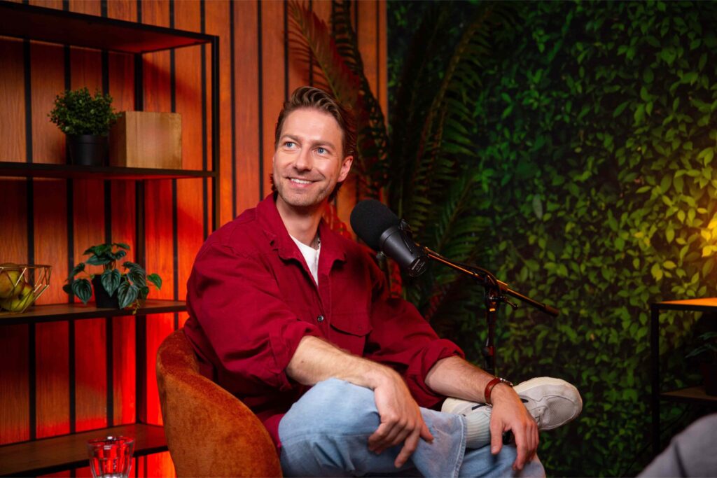 Man in rode blouse zit lachend in een podcaststudio met microfoon en groene planten op de achtergrond.