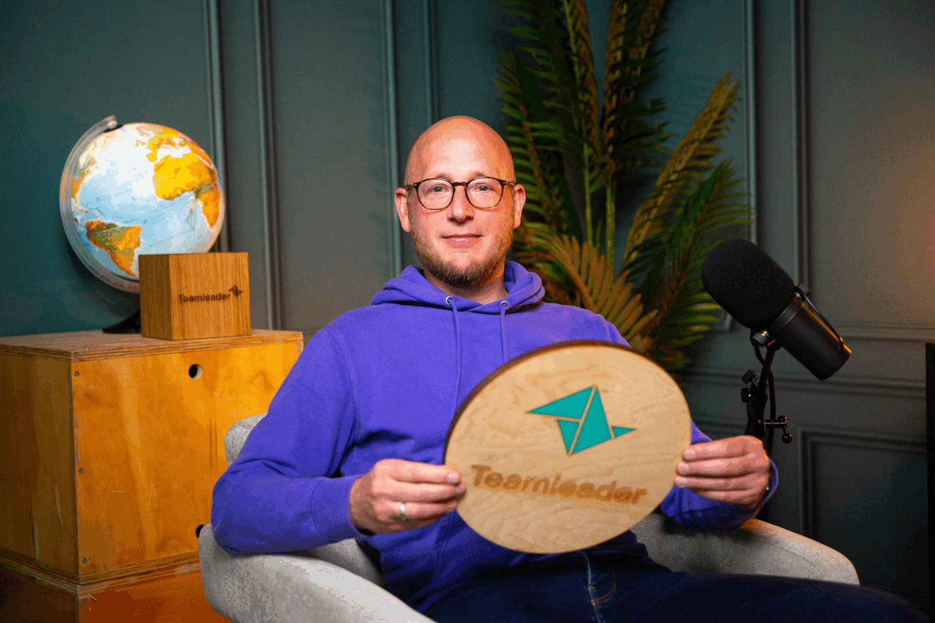 Man in paarse hoodie houdt een houten bord met het Teamleader-logo vast tijdens een opname naast een microfoon.