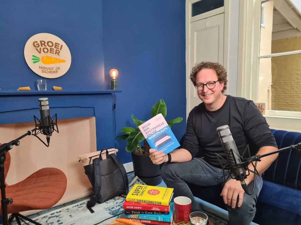 Wout Withagen zit in een podcaststudio en houdt zijn boek "Bouw een Succesvolle Online Marktplaats" omhoog.