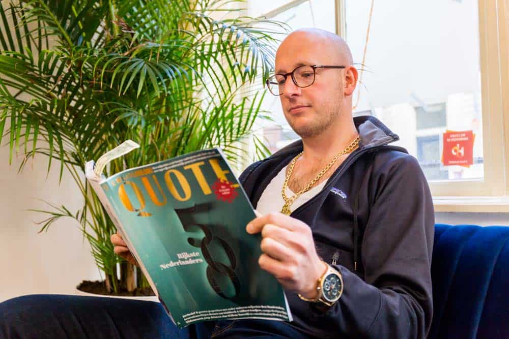 Man met goudkleurige ketting leest de Quote 500 op een bank in een modern interieur.