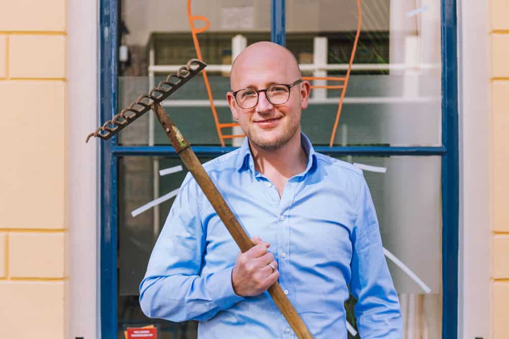 Man in een blauwe blouse poseert met een hark voor een raam met oranje lijntekeningen.